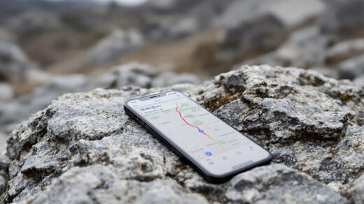 iPhone 18 Pro resting on a rocky outdoor stone surface with rugged terrain blurred in the background, screen showing a Maps navigation interface with a route active, shot at 45-degree angle using a 50mm lens, soft overcast natural light from the left, shallow depth of field, no hands, no people, no text, no watermark, sharp focus, photorealistic, 16:9