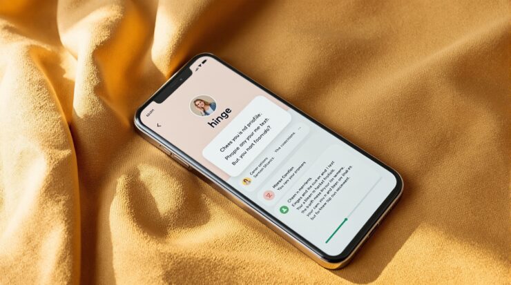 iPhone 15 Pro resting on a warm tan linen surface, Hinge dating app profile visible on screen with a prompt question and text answer partially visible, soft natural window light from the left, 50mm lens, 45-degree angle, no hands, no people, no real profile photos or usernames visible, photorealistic, 16:9