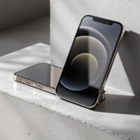 iPhone 18 Pro Max design dummy unit resting upright on its side on a matte warm grey concrete surface, showing the device's thickness profile clearly, shot at 45-degree angle using a 50mm lens, soft natural window light from the left casting long diffused shadows, realistic titanium edge reflections and concrete texture, no hands, no people, no text, no watermark, sharp focus, photorealistic, 16:9